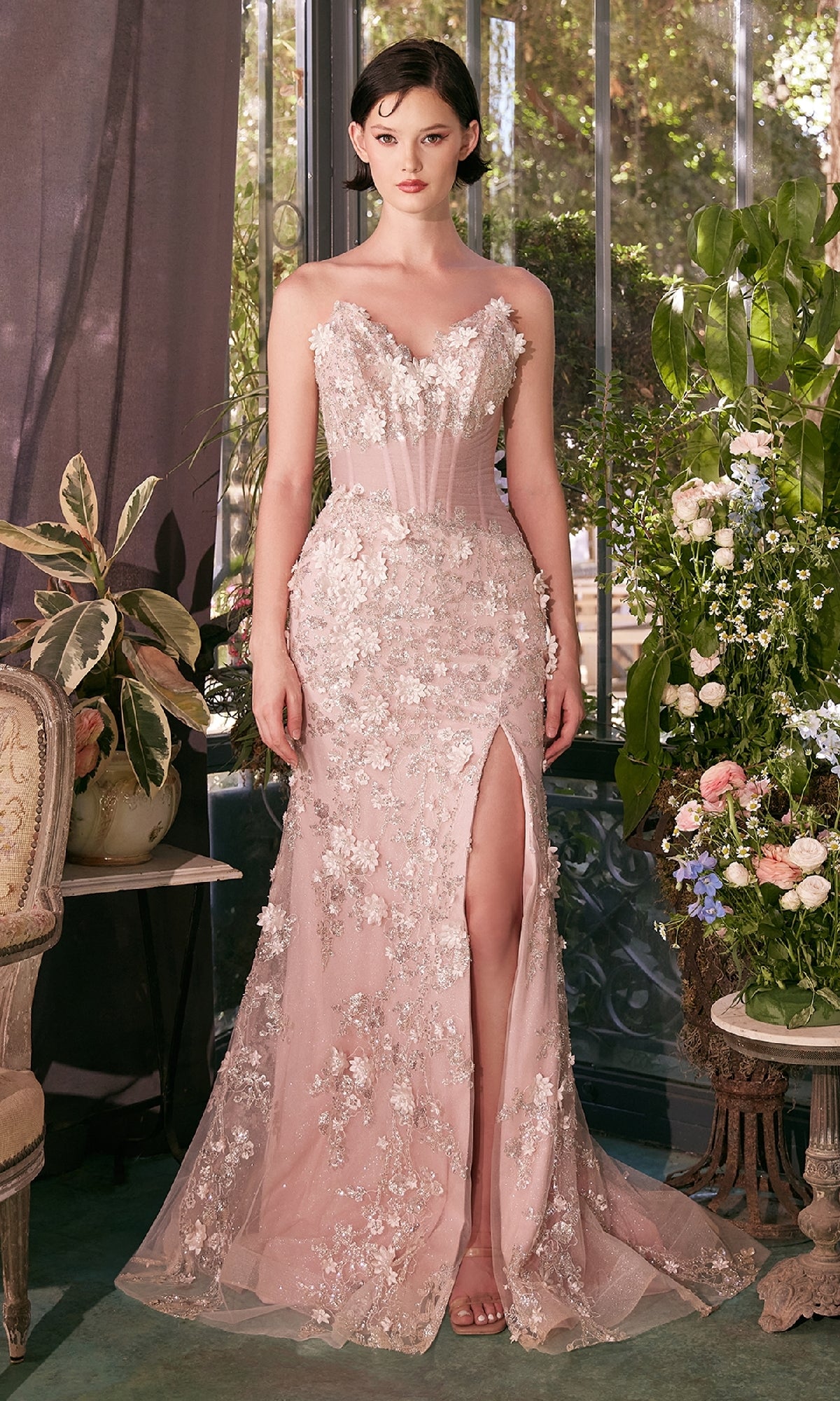 A woman stands indoors wearing the Strapless Floral Prom Gown: Andrea & Leo A1396, a blush pink lace dress with a thigh-high slit; plants and flowers decorate the background.